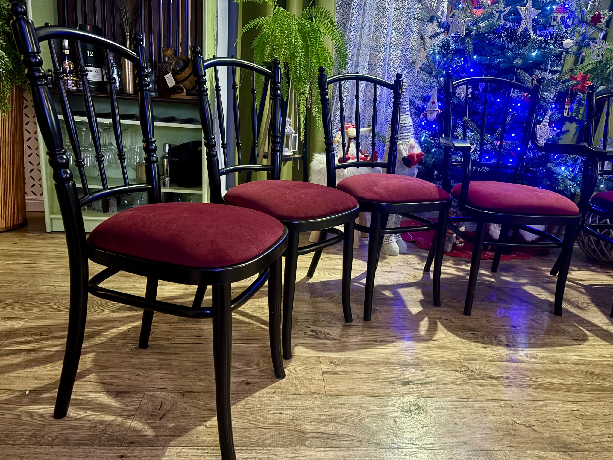 Cztery odnowione, czarne krzesła z bordowymi siedziskami ustawione na drewnianej podłodze, w tle półka z alkoholem i choinka z niebieskimi lampkami.