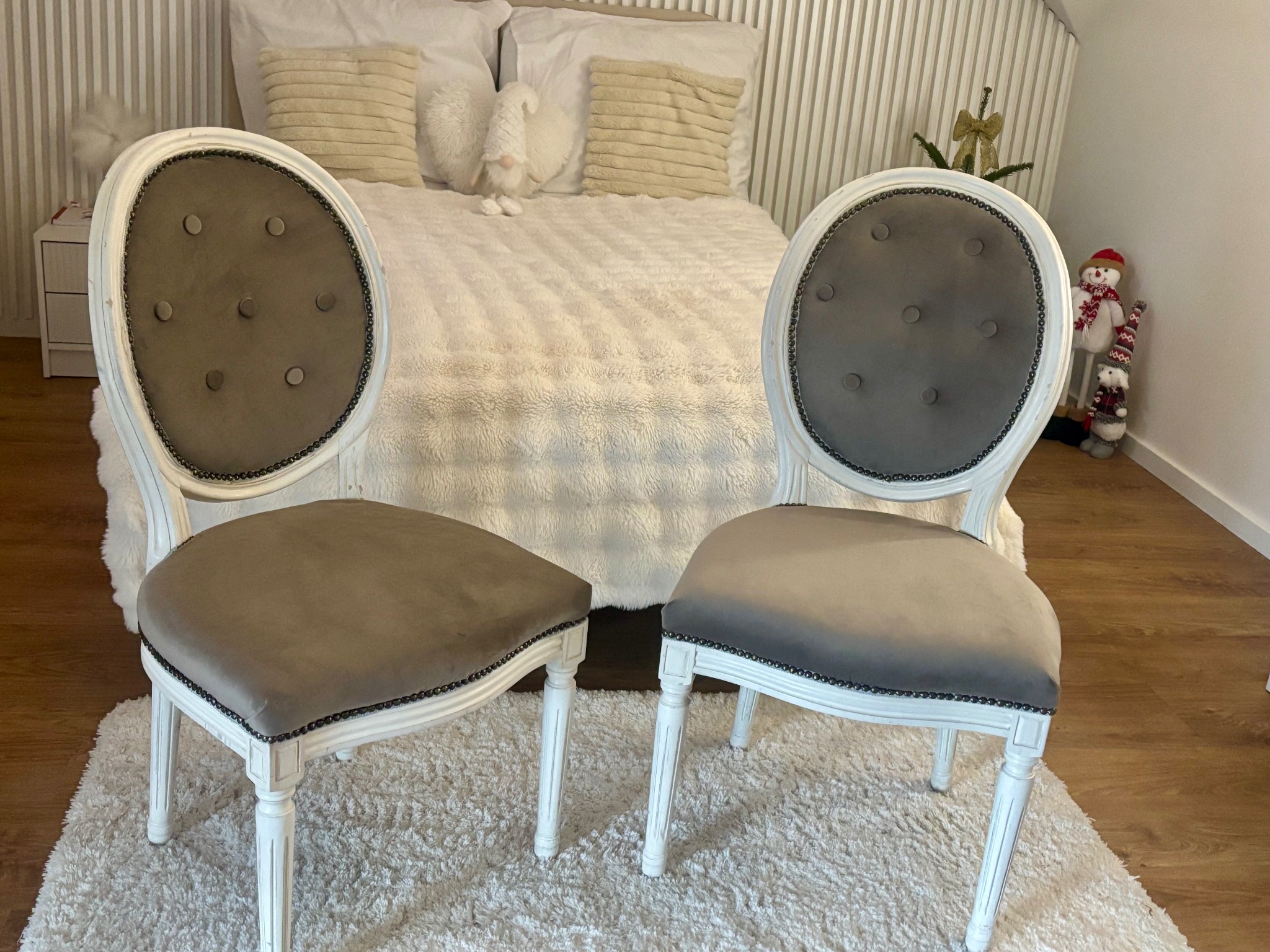 Dwa odnowione, tapicerowane krzesła w stylu Ludwika XVI, z szarym welurem i białymi ramami, ustawione na puszystym białym dywanie, z łóżkiem w tle, tworzące elegancką aranżację wnętrza.