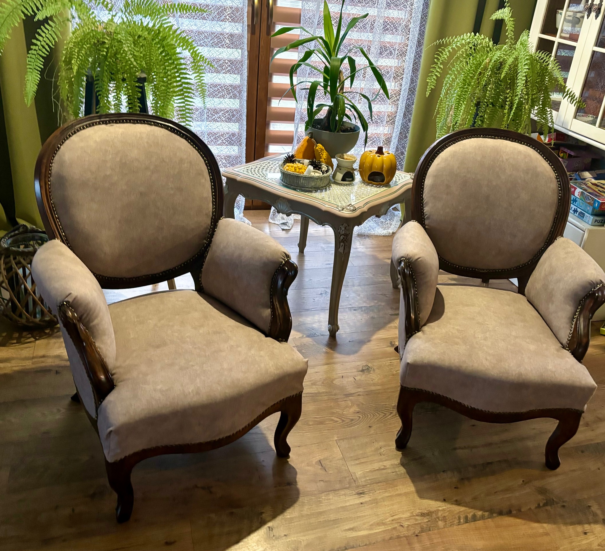Dwa odnowione fotele w stylu vintage z beżową tapicerką i ciemnym drewnianym wykończeniem, ustawione symetrycznie w pomieszczeniu z drewnianą podłogą i stolikiem z dekoracjami.