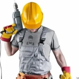 Mężczyzna w ż&oacute;łtym kasku i szarym kombinezonie roboczym trzyma wiertarkę udarową na tle białego studia.