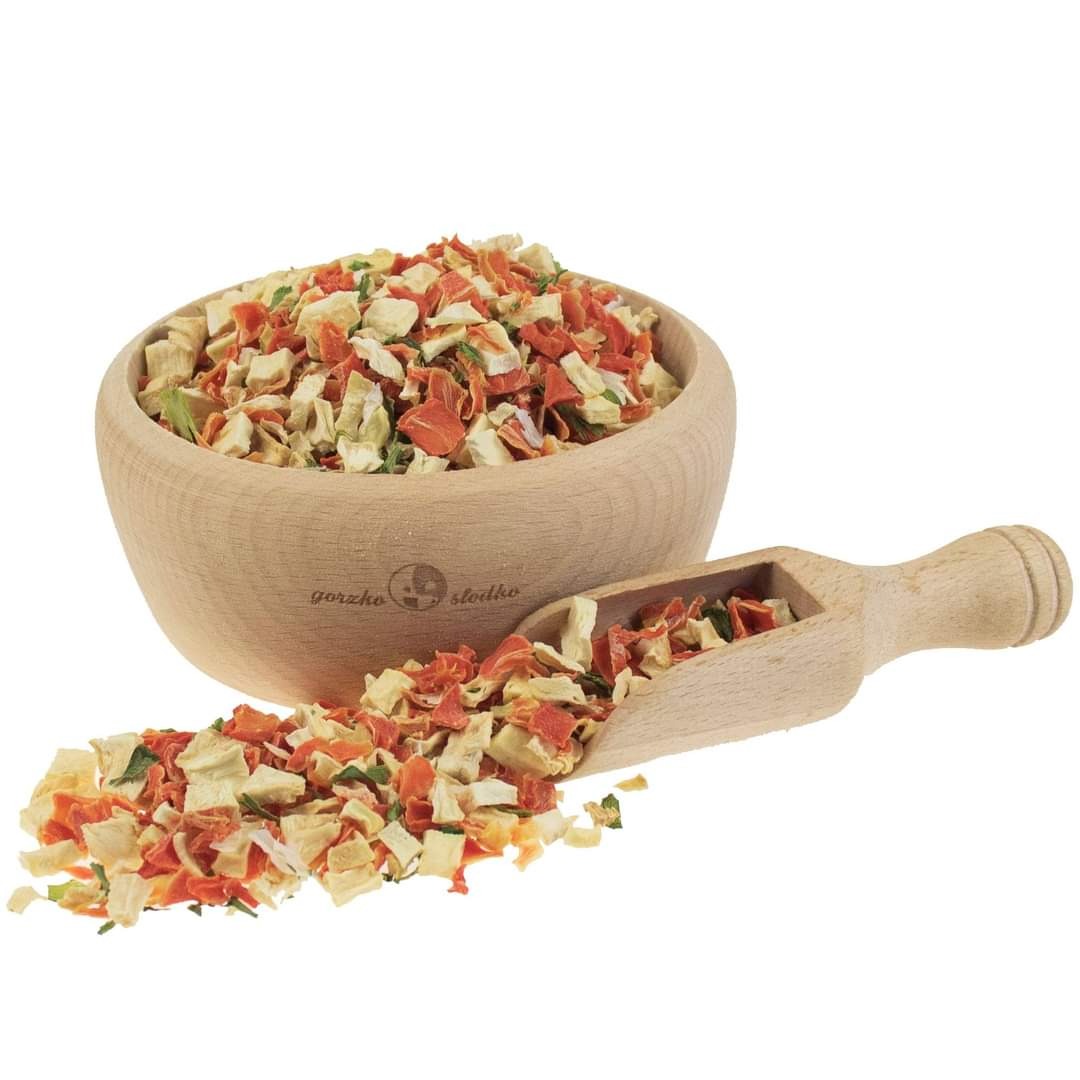 Mieszanka suszonych warzyw (marchew, pietruszka, seler) w drewnianej miseczce z logo 'gorzko słodko' i drewnianej łopatce na białym tle.