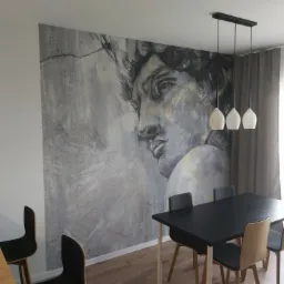Jadalnia z dużą fototapetą przedstawiającą popiersie Dawida w odcieniach szarości i bieli, wisząca lampa z trzema białymi kloszami, czarny stół i krzesła w stylu skandynawskim.