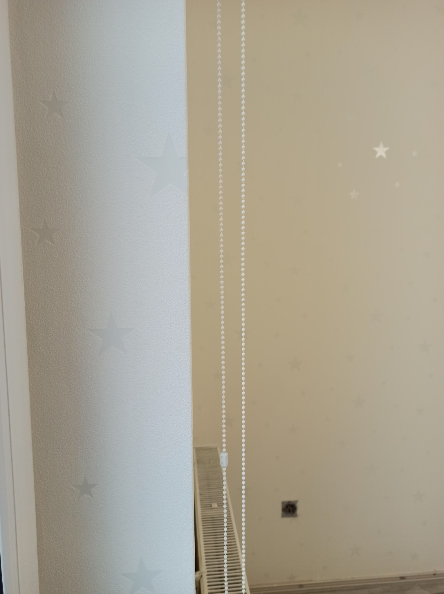 Jasna ściana z delikatnym wzorem gwiazd, częściowo zakryta przez pionową kolumnę również w gwiazdy i sznurkowy mechanizm rolety, widoczny fragment grzejnika.
