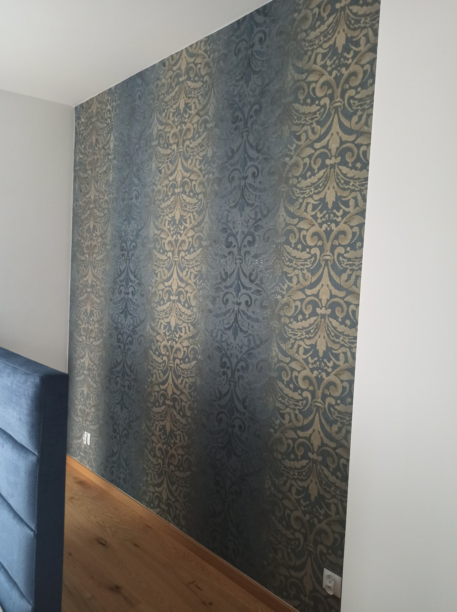 Ściana z wzorzystą tapetą w odcieniach szarości i złota, fragment niebieskiego zagłówka łóżka, drewniana podłoga i biała ściana boczna.