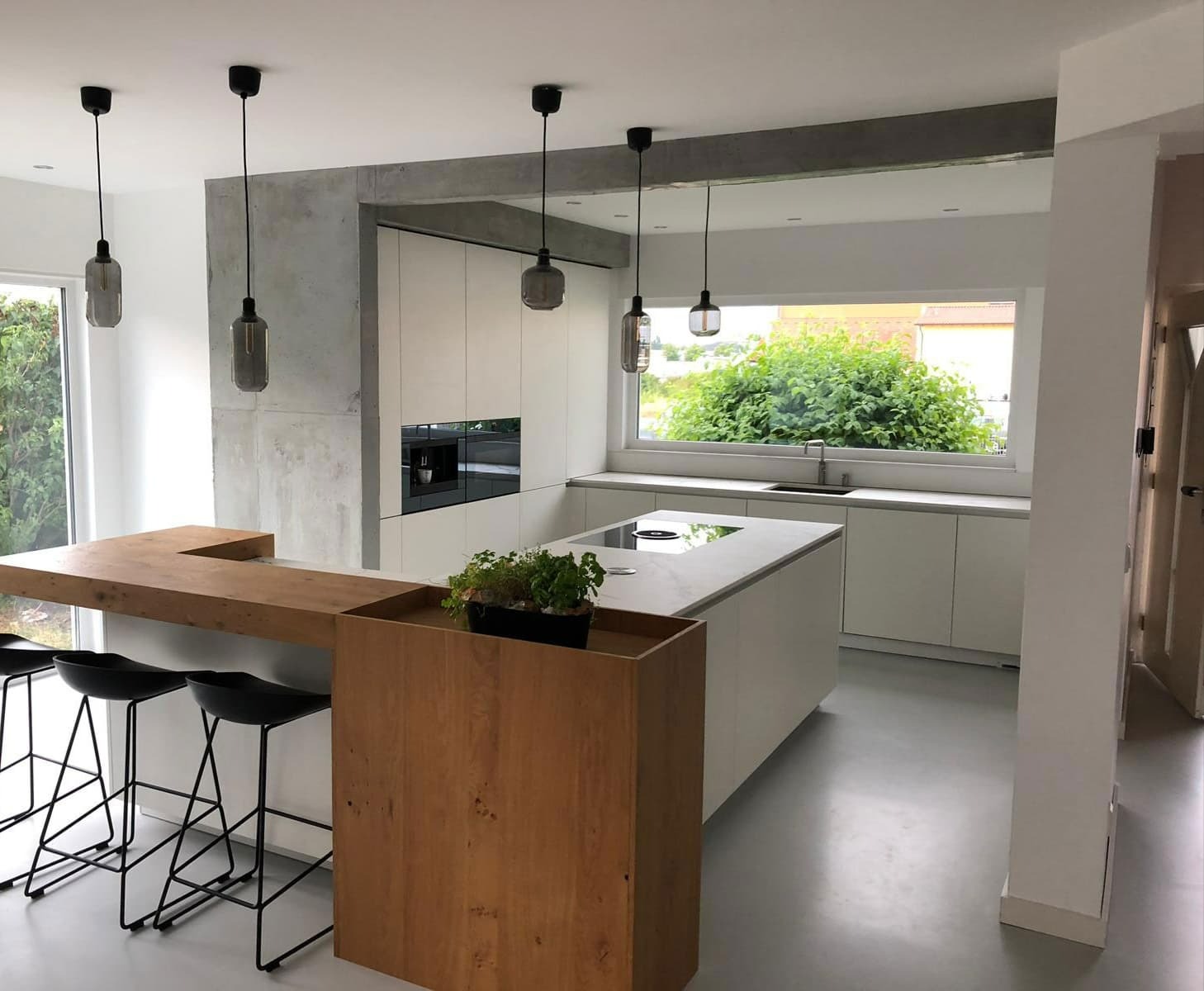 Nowoczesna kuchnia z wyspą i drewnianym barkiem, minimalistyczne białe szafki z wbudowanym ekspresem do kawy, betonowe elementy architektoniczne, szare podłogi i wiszące lampy w szklanych kloszach.