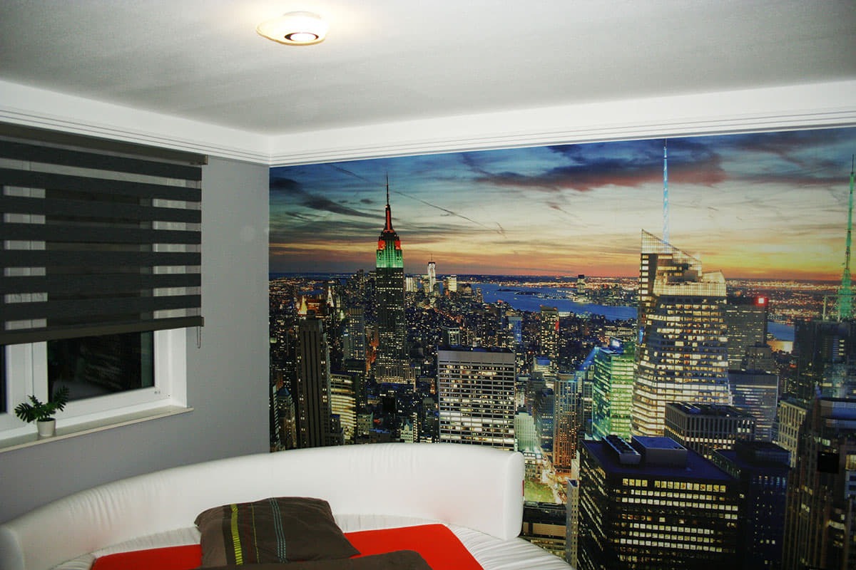 Fototapeta przedstawiająca panoramę Nowego Jorku o zmierzchu, z widocznym Empire State Building, zamontowana na ścianie pokoju z oknem przesłoniętym roletami i zaokrąglonym zagłówkiem łóżka.