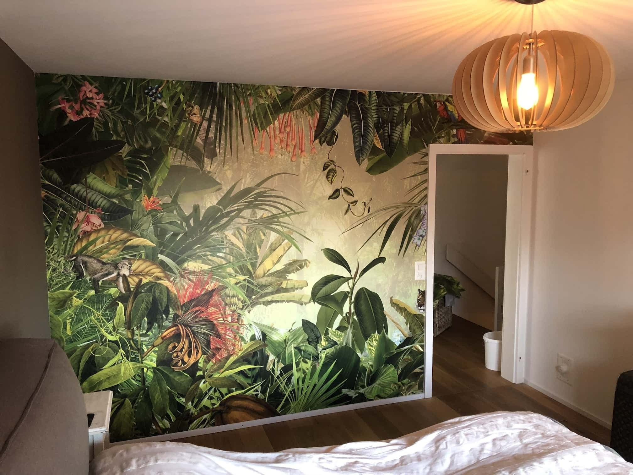 Ściana w sypialni z fototapetą przedstawiającą motyw dżungli, widoczny fragment łóżka z białą pościelą, na suficie drewniana lampa wisząca, w tle otwarte drzwi do innego pomieszczenia.