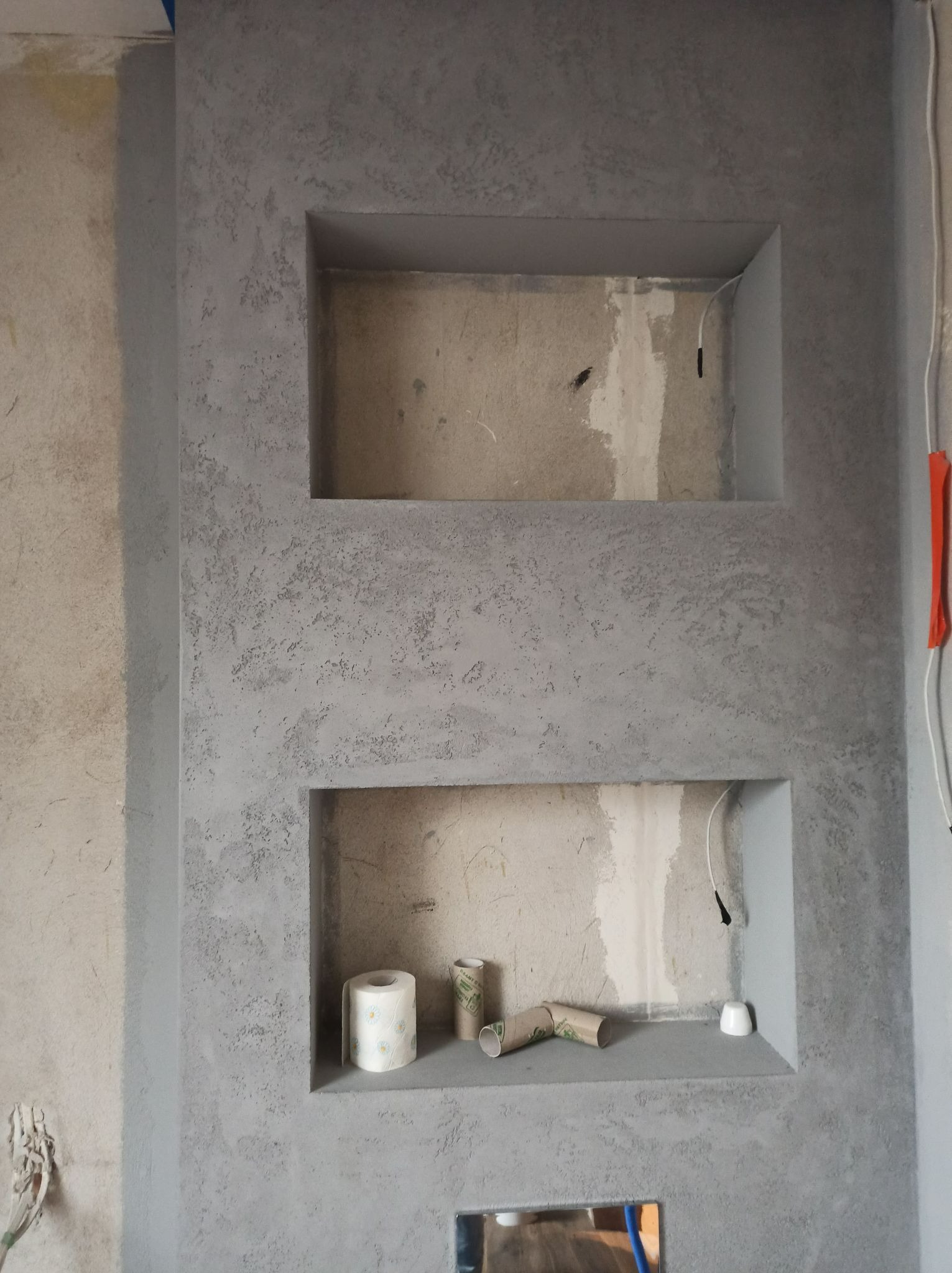 Ściana z szarą, dekoracyjną strukturą i wnękami, w jednej z nich rolka papieru toaletowego i puste rolki po papierze, widoczne kable elektryczne.