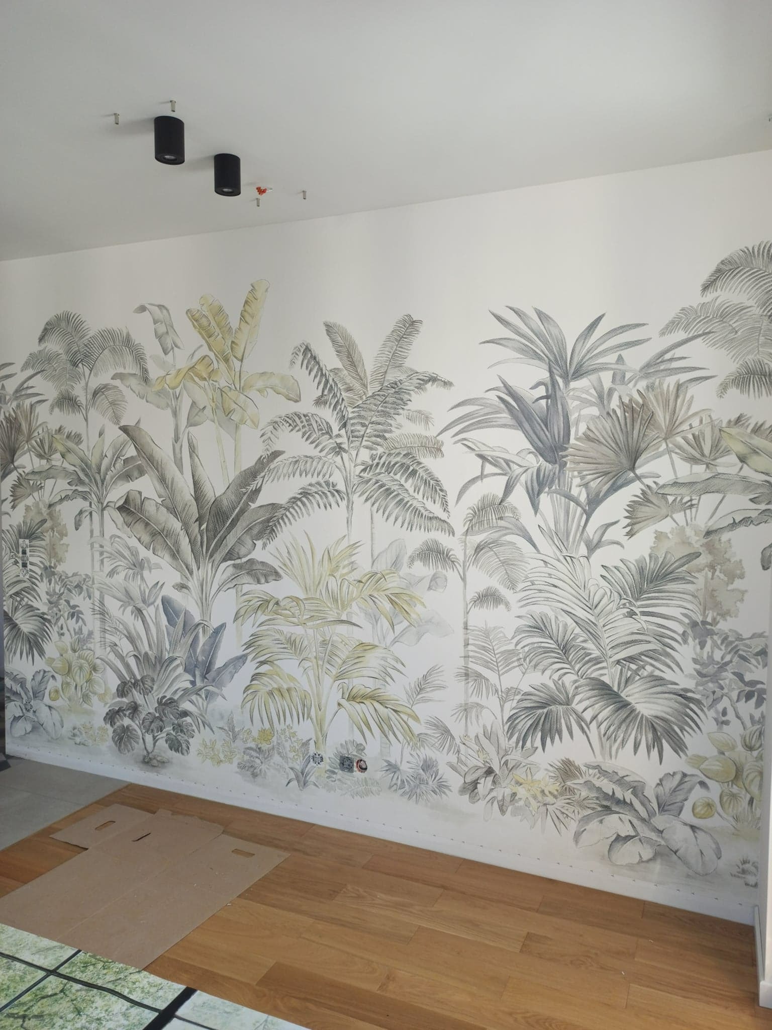 Ściana z tapetą w motywy tropikalne, widoczne gniazdka elektryczne i fragment podłogi z paneli oraz karton ochronny.