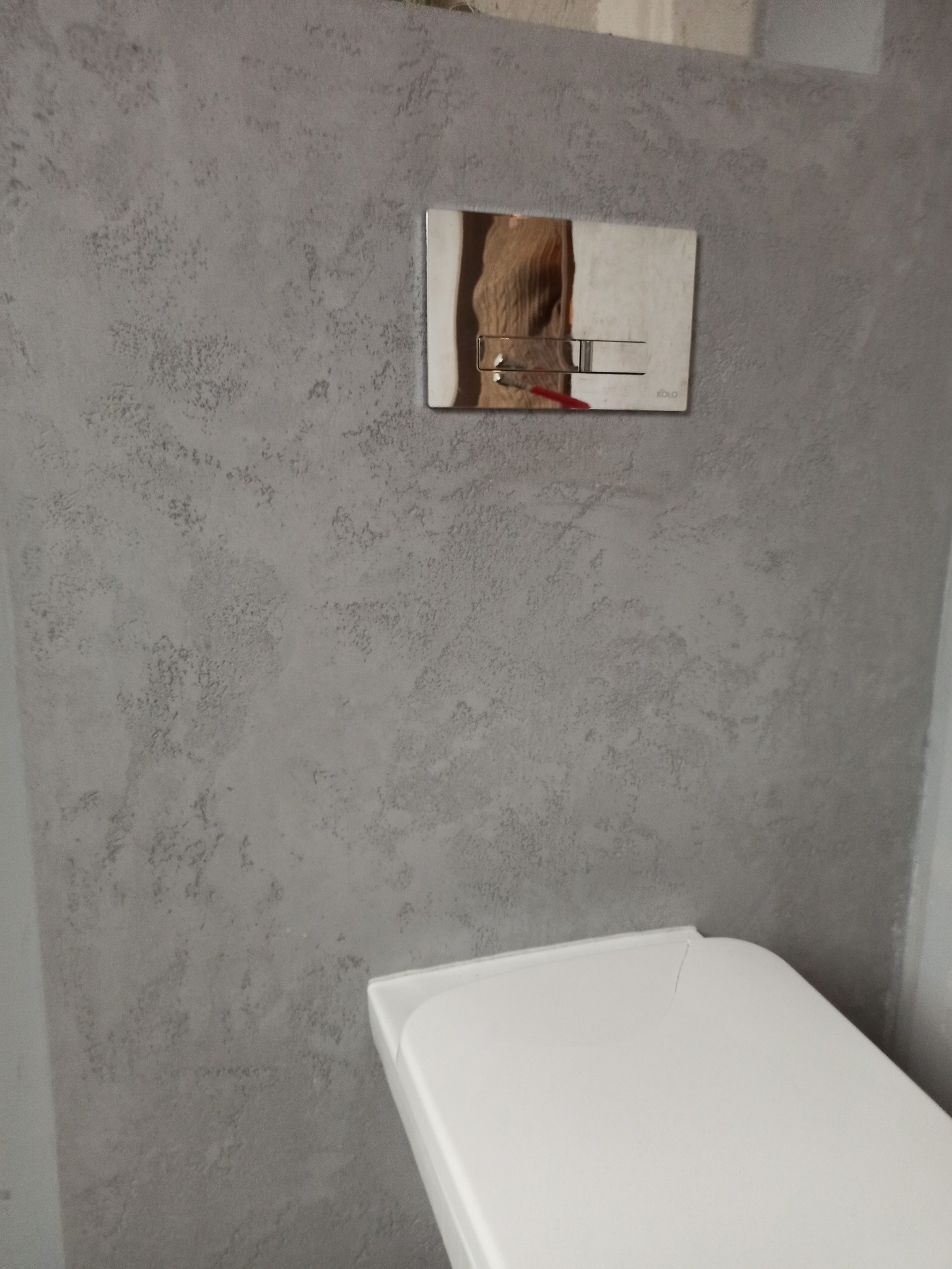 Szara ściana w łazience z dekoracyjną fakturą, chromowany panel spłuczki toalety marki Koło, widoczny fragment białej toalety.