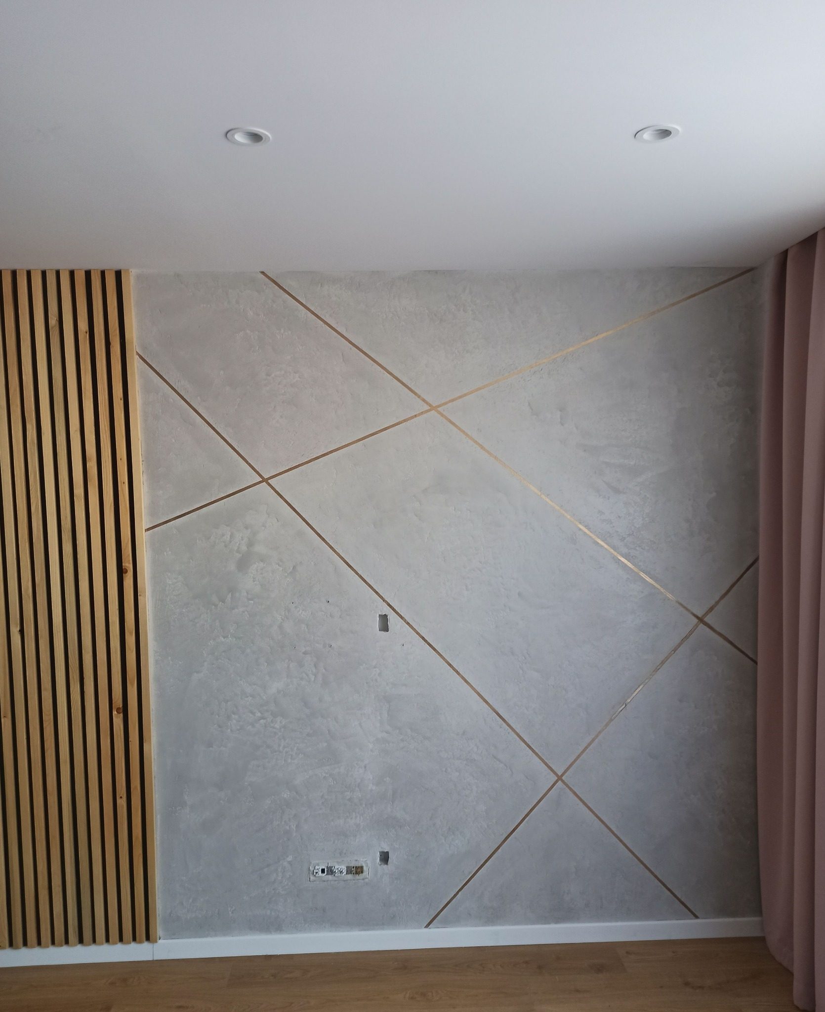 Ściana z szarą gładzią dekoracyjną i złotymi paskami tworzącymi geometryczny wzór, drewniane lamele po lewej stronie, widoczne gniazdka elektryczne i fragment zasłony.