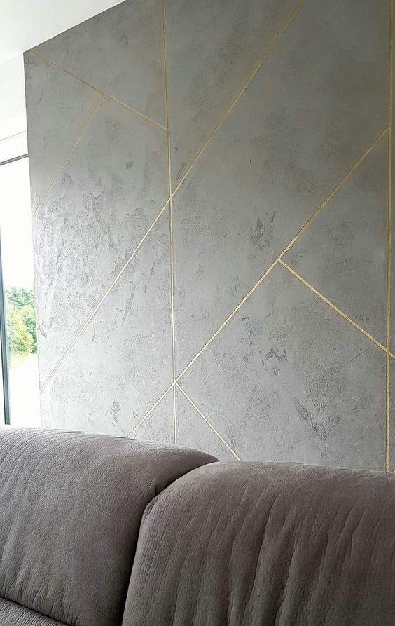 Szara ściana z dekoracyjną fakturą i złotymi liniami tworzącymi geometryczny wzór, widoczny fragment sofy w dolnej części kadru, okno z widokiem na zieleń w lewym górnym rogu.