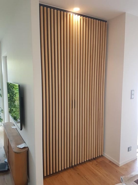 Drewniana ściana z pionowych listew z czarną ramą na górze, oświetlona punktowym światłem, na tle parkietu i białej ściany z włącznikiem.