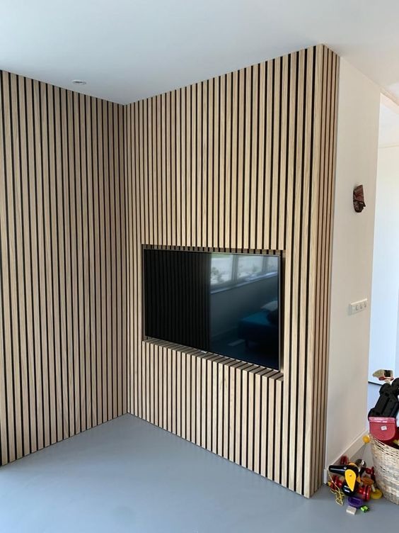 Ściana wykończona panelami lamelowymi z wnęką na telewizor, widok na szare panele podłogowe i fragment pokoju dziecięcego z koszem na zabawki.