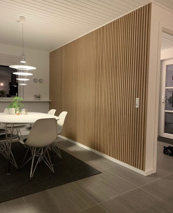 Ściana w jadalni wykończona panelami lamelowymi w odcieniach beżu i brązu, widoczny fragment białego sufitu z lampą wiszącą, biały stół z krzesłami i szary dywan.