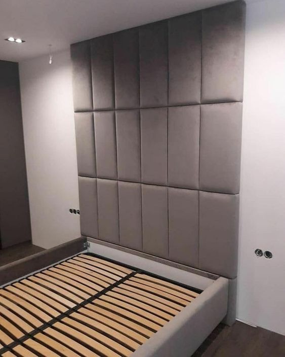 Tapicerowany zagłówek łóżka w odcieniach szarości, składający się z pionowych paneli, zamontowany na ścianie nad drewnianym stelażem łóżka.