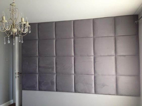 Ściana sypialni wykończona panelami tapicerowanymi w odcieniach szarości, nad białym zagłówkiem łóżka, z dekoracyjnym żyrandolem kryształowym w rogu.