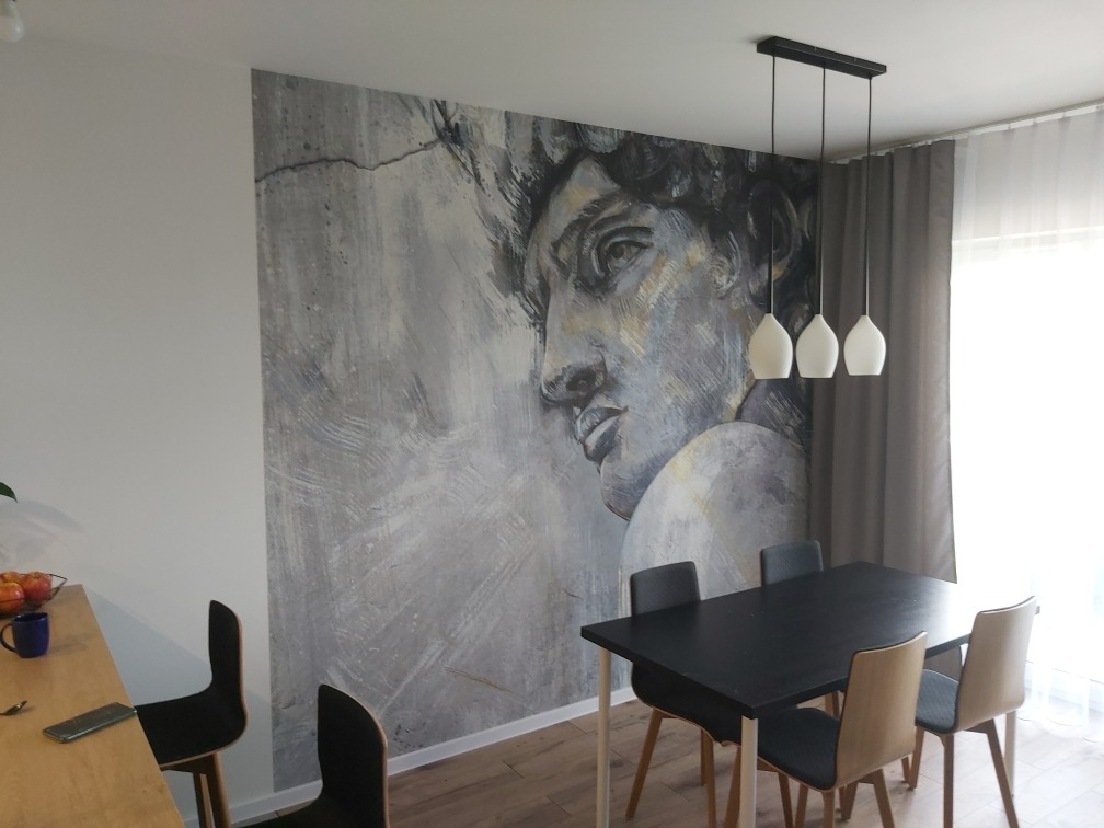 Jadalnia z dużą fototapetą przedstawiającą popiersie Dawida w odcieniach szarości i bieli, wisząca lampa z trzema białymi kloszami, czarny stół i krzesła w stylu skandynawskim.