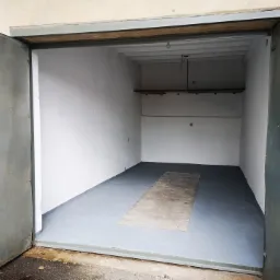 Wnętrze pustego, odświeżonego magazynu z szarą podłogą, białymi ścianami i otwartymi, stalowymi drzwiami na zewnątrz.