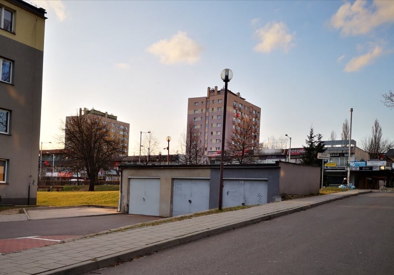 Szereg garaży blaszanych w zabudowie szeregowej na tle bloków mieszkalnych i błękitnego nieba o zmierzchu.
