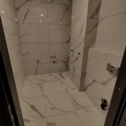 Wykończona łazienka z białymi płytkami imitującymi marmur na ścianach i podłodze, widoczne przyłącza hydrauliczne i fragment czarnej ramy drzwi.