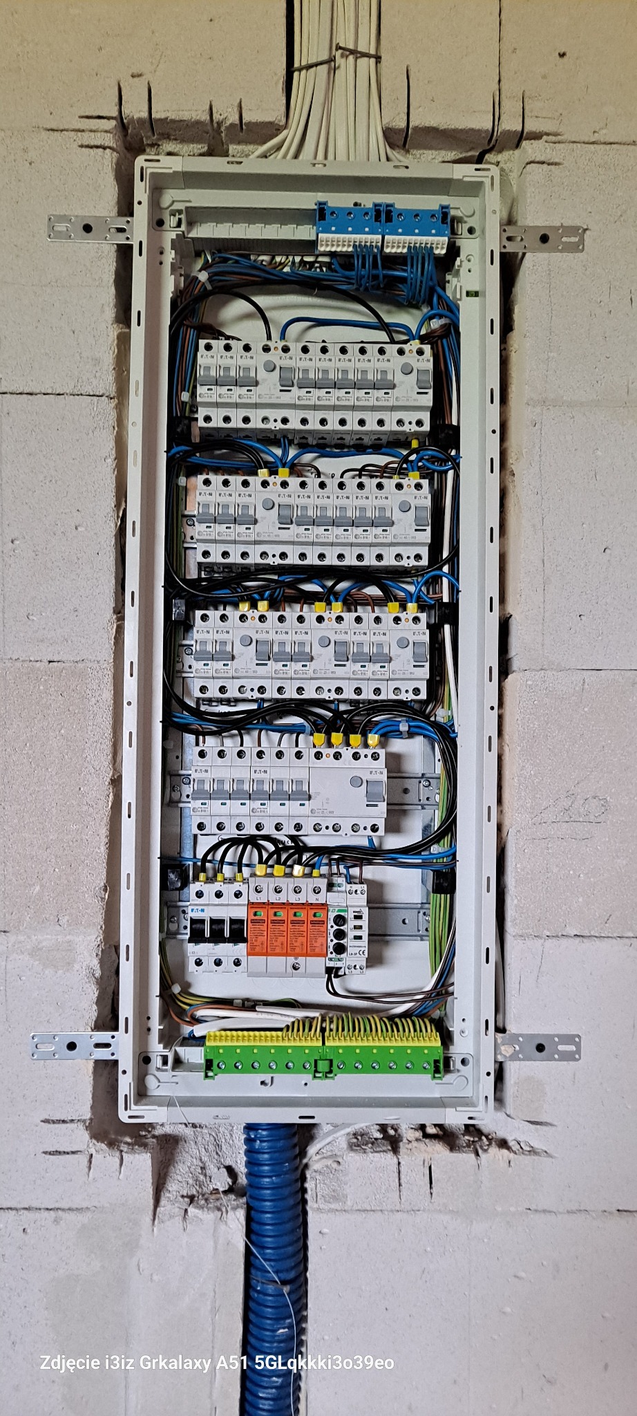 Rozdzielnica elektryczna z zainstalowanymi bezpiecznikami, zabezpieczeniami przepięciowymi i listwami zaciskowymi, zamontowana na surowej ścianie z betonu komórkowego, z widoczną niebieską rurą...