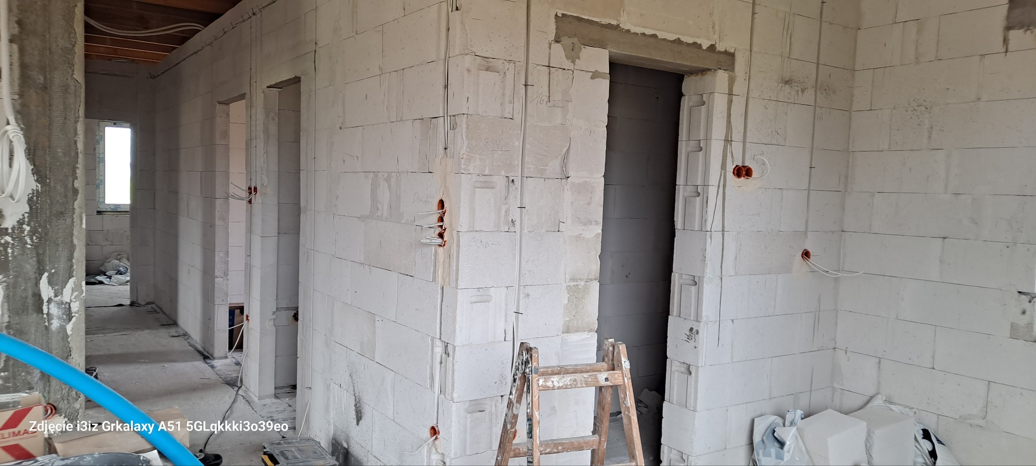 Wnętrze w trakcie budowy z białych bloczków, widoczne puszki elektryczne w ścianach i drabina oparta o framugę drzwiową.