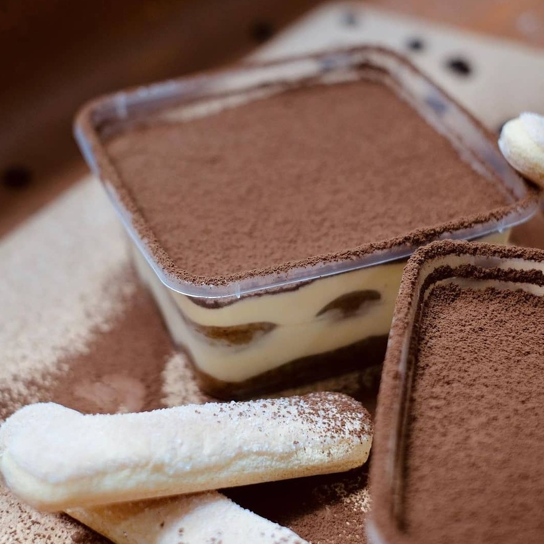 Tiramisu w przezroczystym plastikowym pojemniku, posypane kakao, obok biszkopty z cukrem pudrem, całość na drewnianym tle z rozsypanym kakao.