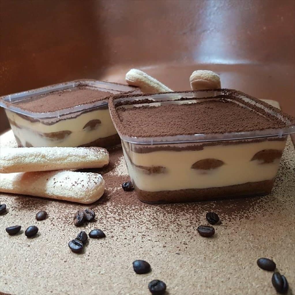 Dwa pojemniki z Tiramisu Classico posypane kakao, obok biszkopty Savoiardi i ziarna kawy na brązowym tle.