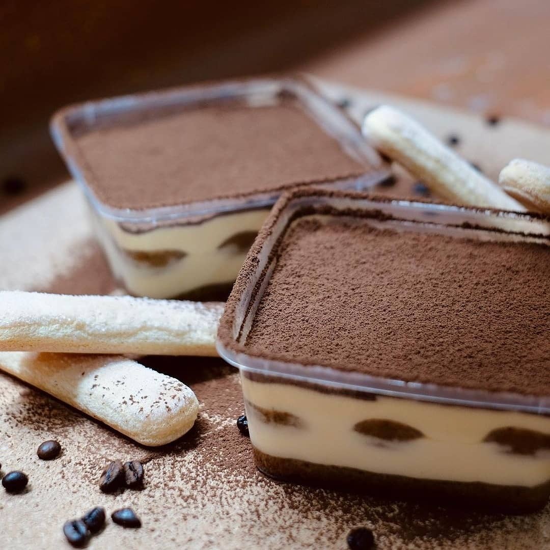 Dwa porcje Tiramisu Classico w przezroczystych pojemnikach, posypane kakao, ułożone na pudrowanym blacie z biszkoptami i ziarnami kawy.