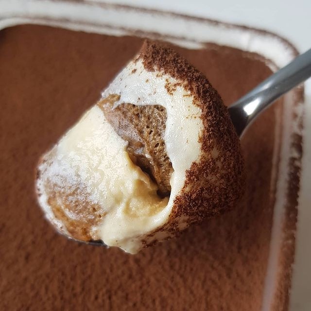 Kremowe Tiramisu Classico na łyżce, posypane kakao, z widocznymi warstwami kremu i biszkoptów nasączonych kawą.