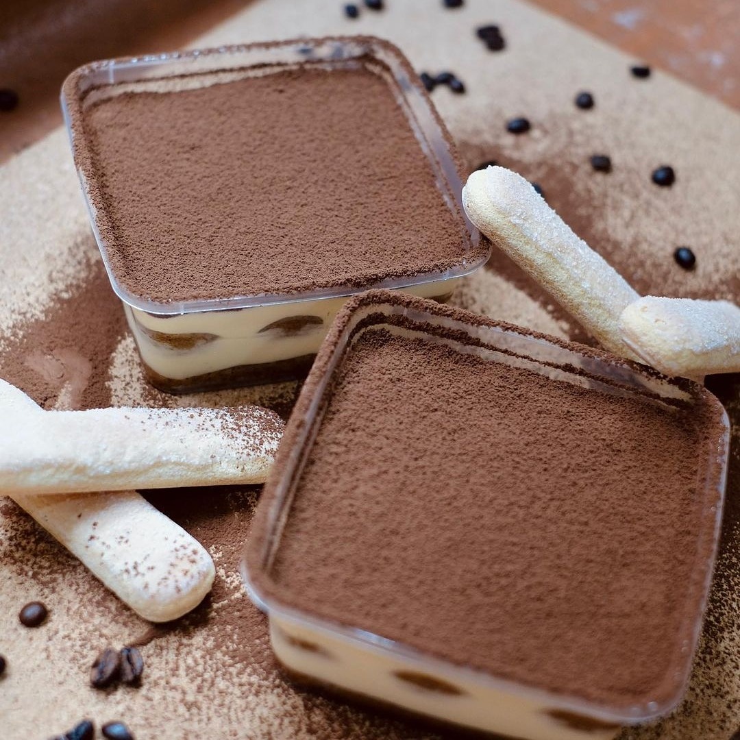 Dwa porcje Tiramisu Classico w przezroczystych, kwadratowych pojemnikach, posypane kakao, obok biszkopty Savoiardi i rozsypane ziarna kawy na posypanym kakao blacie.