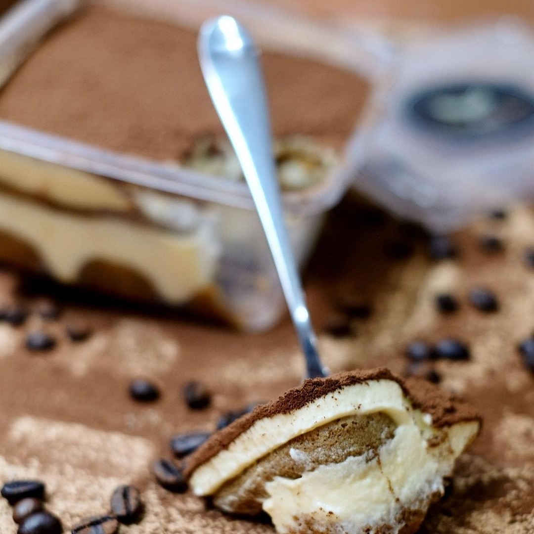 Kawałek Tiramisu Classico na brązowym tle z rozsypanymi ziarnami kawy, obok pojemnik z deserem i łyżeczka.