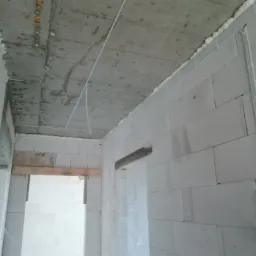 Wnętrze w stanie surowym: widoczne przewody elektryczne w peszlach przy suficie i ścianie z betonu komórkowego, zabezpieczone pianką montażową, z fragmentami wystających kabli.