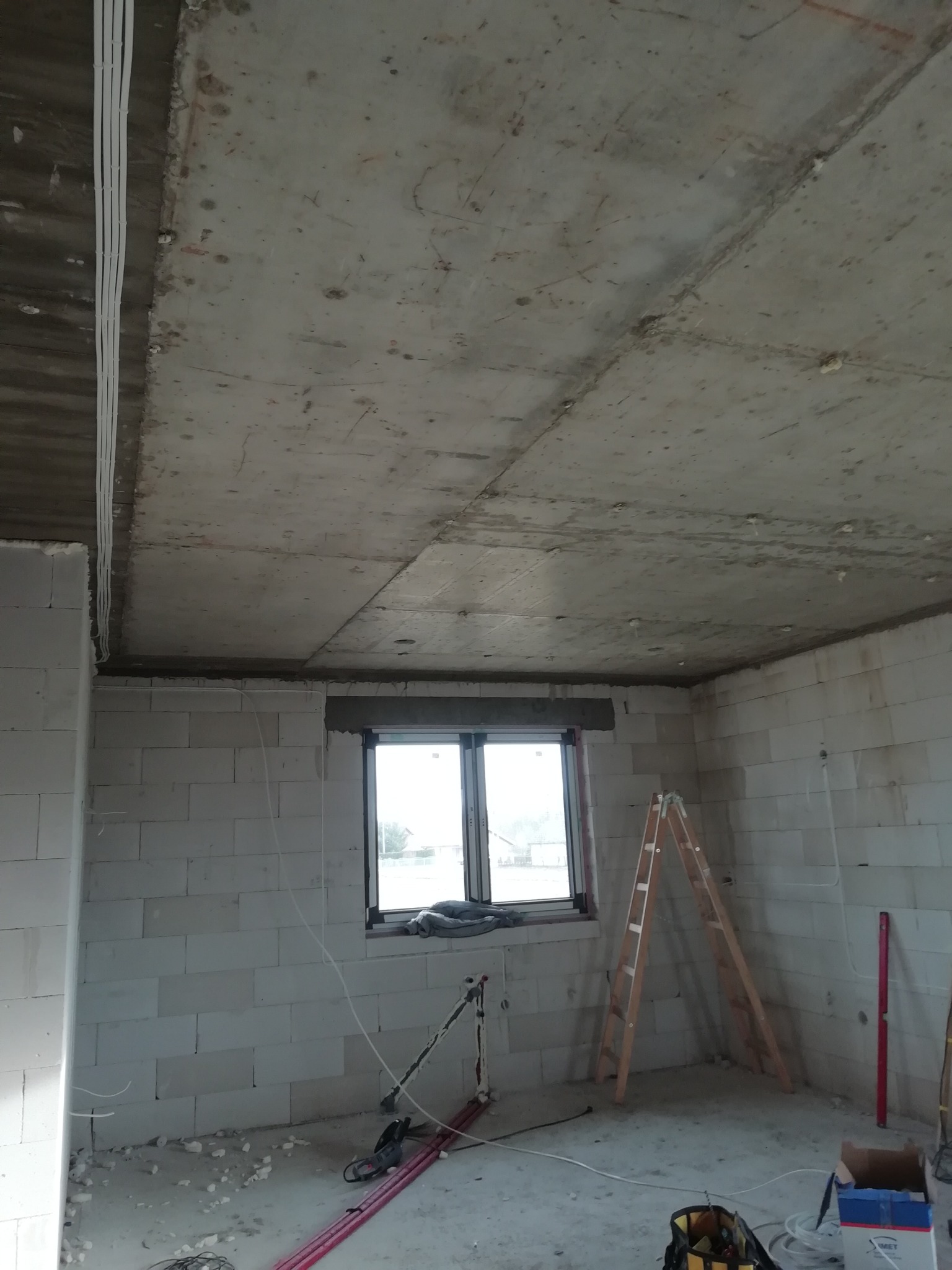Surowy stan wnętrza z widoczną instalacją elektryczną w peszlach pod sufitem, ściany z białych bloczków, okno z roletą, drabina, poziomica, narzędzia i przewody na podłodze.