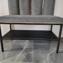 Szary tapicerowany puf z metalową konstrukcją i złotymi ozdobnymi nitami, stojący na marmurowej podłodze.