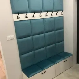 Turkusowy panel ścienny z tapicerowanymi kwadratami, wbudowanym siedziskiem z trzema szafkami i rzędem czarnych haczyków na białej listwie, zamontowany w korytarzu.