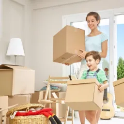 Rodzina z dwójką dzieci przenosi kartony w jasnym pokoju z kominkiem i wyjściem na taras.