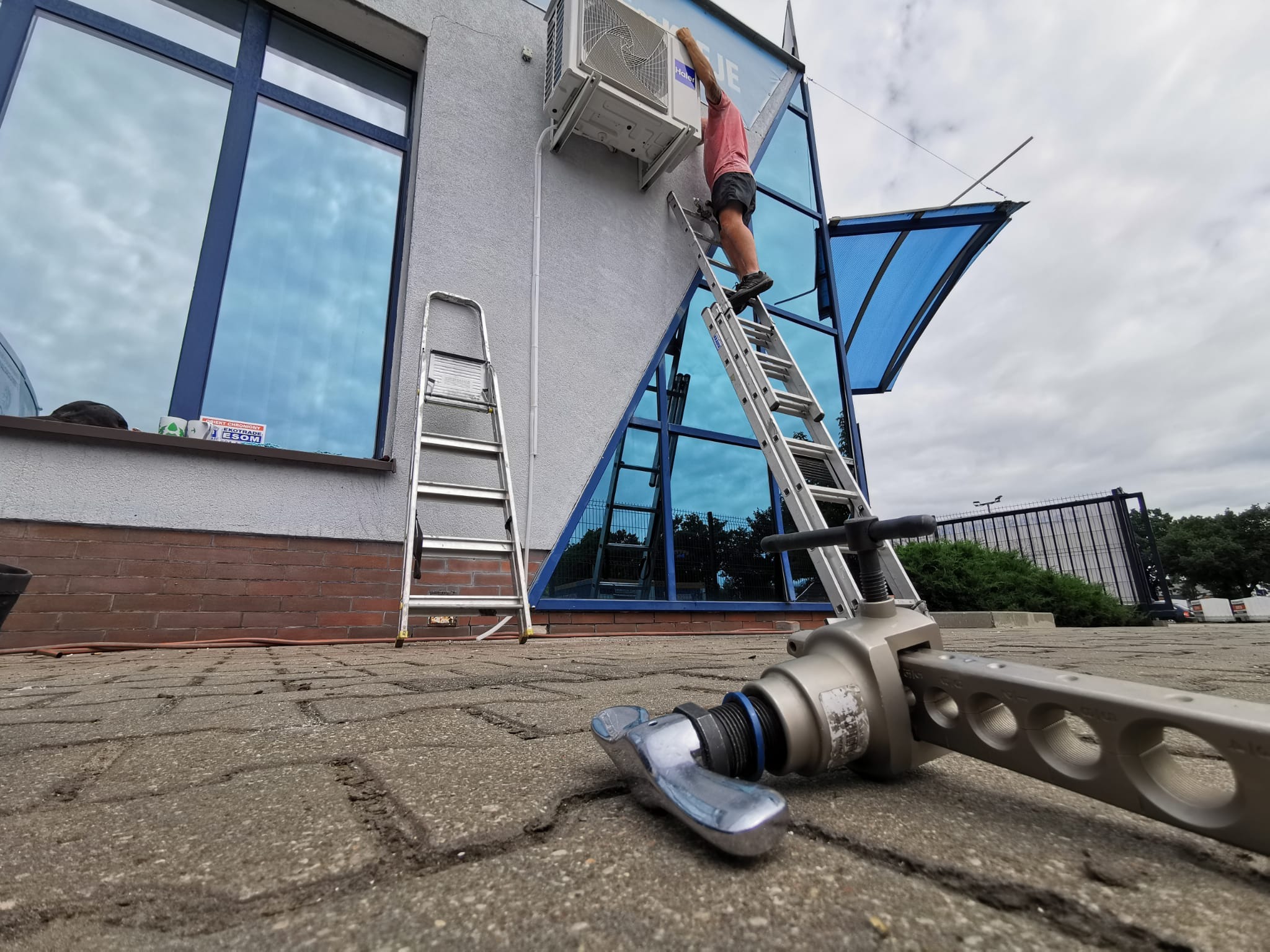 Monteraż jednostki zewnętrznej klimatyzatora na elewacji budynku z niebieskimi oknami, widoczne dwie drabiny i narzędzie do montażu na pierwszym planie.