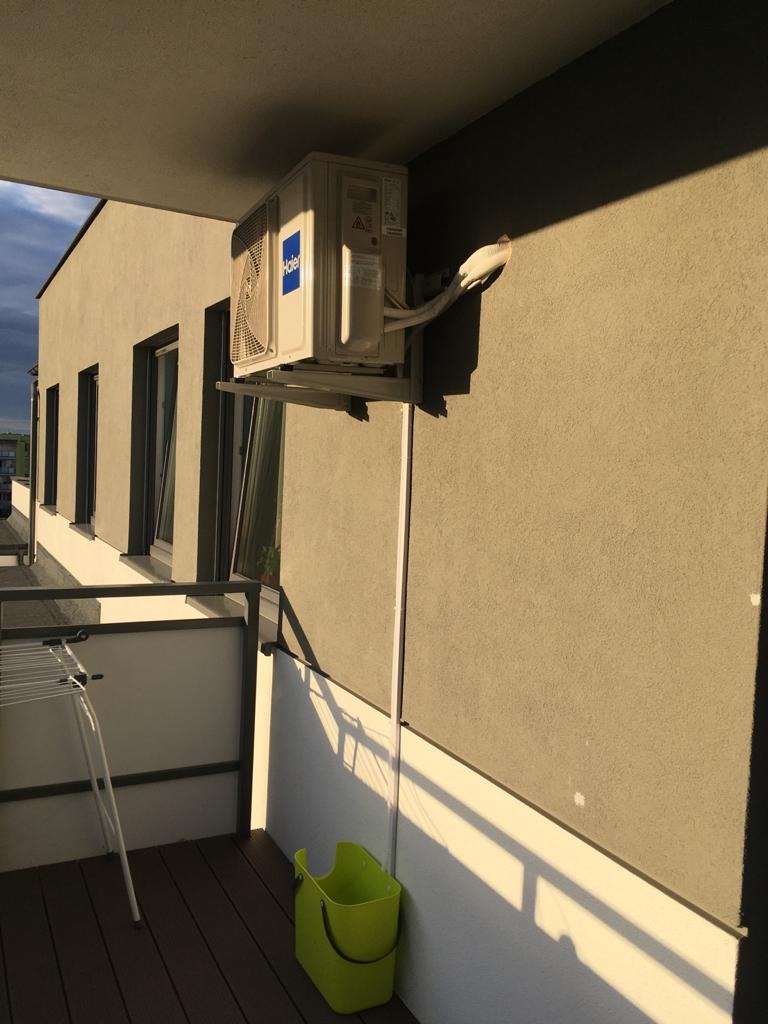 Zewnętrzna jednostka klimatyzacji Haier zamontowana na balkonie, z widocznym odpływem skroplin i zielonym koszem pod spodem, oświetlona popołudniowym słońcem.