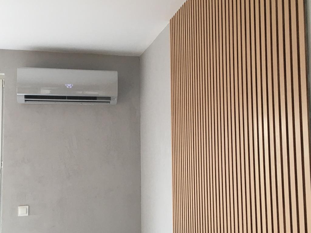 Biała jednostka wewnętrzna klimatyzacji zamontowana na szarej ścianie z dekoracyjną drewnianą ścianką działową z pionowych listew po prawej stronie.