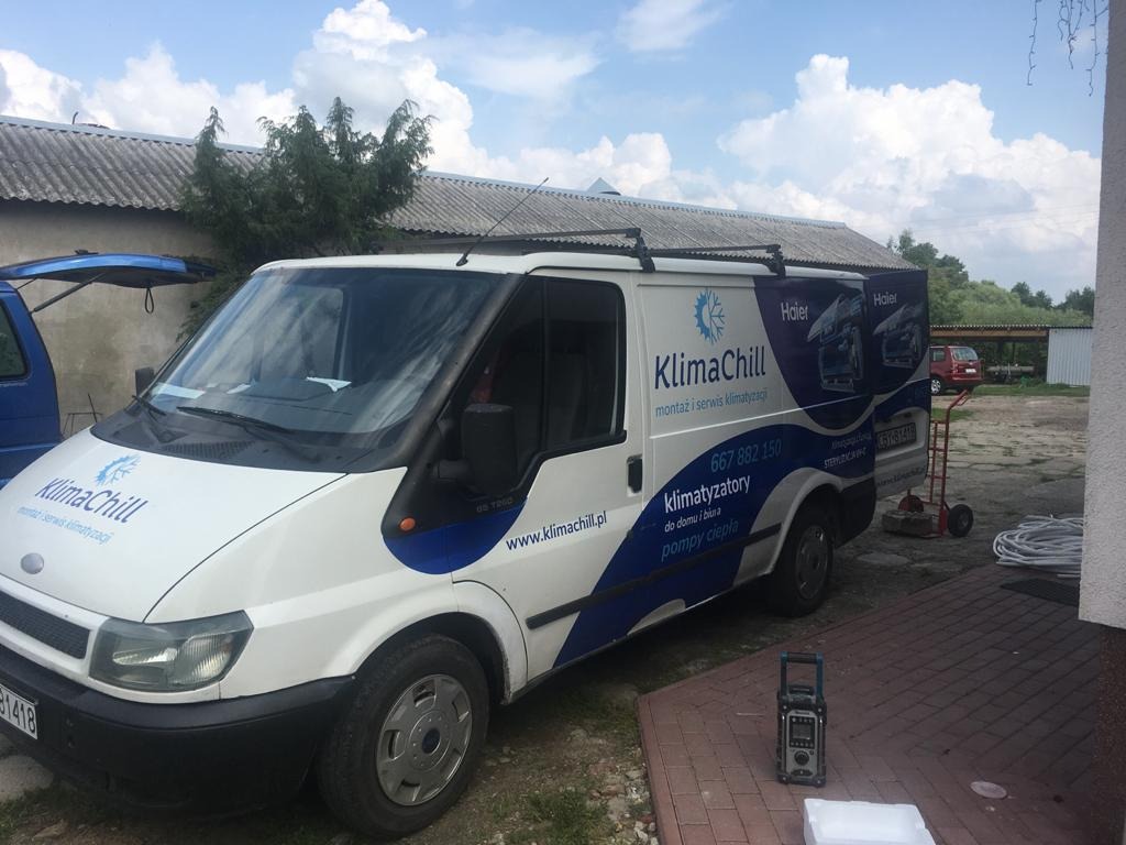 Biały samochód dostawczy z logo firmy KlimaChill, zaparkowany na podwórku z kostki brukowej, z widocznym numerem telefonu i adresem strony internetowej. Na samochodzie reklama klimatyzatorów Haier.