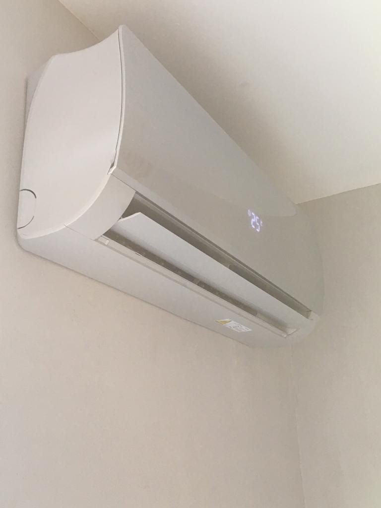 Biały klimatyzator ścienny z wyświetlaną temperaturą 25 stopni Celsjusza, zamontowany w rogu pokoju na beżowej ścianie.