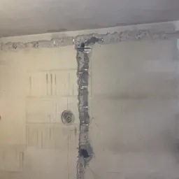 Ściana w trakcie remontu z wyżłobionymi bruzdami na przewody elektryczne, widoczne metalowe uchwyty mocujące kable, fragment osoby w lewym dolnym rogu.