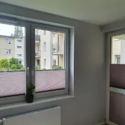 Wnętrze pokoju z dwoma oknami i drzwiami balkonowymi, wszystkie osłonięte plisowanymi roletami w odcieniach brązu, na parapecie okna stoi mała roślina doniczkowa, widok na budynki mieszkalne za oknem.