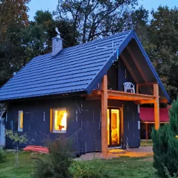 Niedokończony dom murowany z czarną elewacją, widoczne ocieplenie, drewniane elementy konstrukcyjne werandy i drabina oparta o ścianę, wieczorne oświetlenie wnętrza.