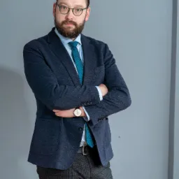 Portret mężczyzny z brodą w okularach, ubrany w niebieską marynarkę i wzorzysty krawat, pozującego na szarym tle ze skrzyżowanymi rękami.