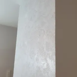 Ściana pokryta dekoracyjną, srebrzystą fakturą, widoczna z bliska, z delikatnym połyskiem i subtelnymi nierównościami, sąsiadująca z gładką, beżową ścianą.