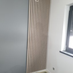 Illia Ivanov wykończenie wnętrz - Montaż pionowych, drewnianych lameli dekoracyjnych na szarej ścianie w rogu pokoju, widoczne elementy mocujące i zabezpieczona folią podłoga.