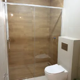 Nowoczesna łazienka z kabiną prysznicową ze szklanymi przesuwnymi drzwiami, podwieszaną toaletą z białym stelażem i płytkami imitującymi drewno na ścianach wewnątrz kabiny.